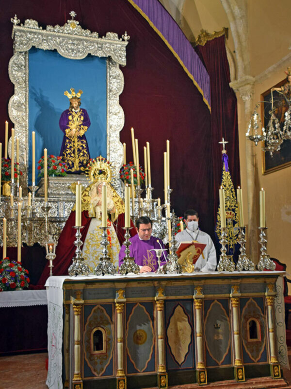 Un altar en una iglesia, con una estatua dorada de un santo en el centro. A la izquierda hay flores y velas encendidas, mientras que a la derecha se observa un vitral con una imagen de Cristo. La estatua está rodeada por velas y adornos ornamentales, todo en un contexto religioso.