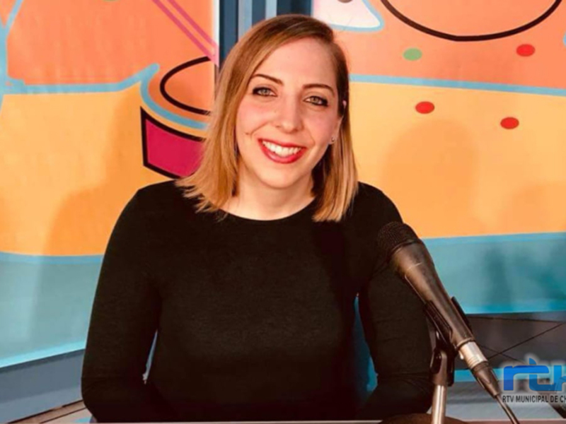 A una persona sonriendo frente a un fondo colorido con elementos gráficos. La persona está sentada en una silla y tiene un micrófono frente a ella, lo que sugiere que está en un entorno de radio o podcast. La imagen es parte del contenido de RTVC, una emisora municipal de Chipiona.