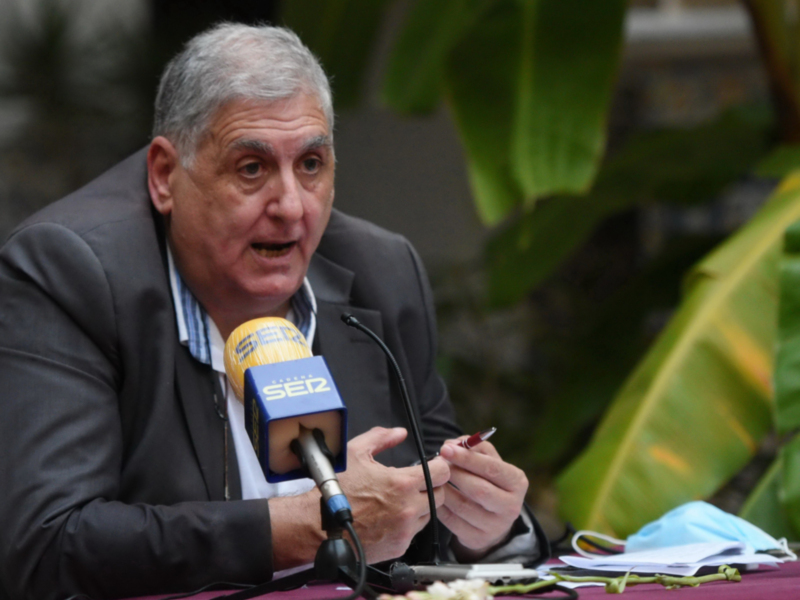 El hombre está hablando en un micrófono con una etiqueta "SE12" mientras sostiene un cigarrillo. La imagen está rodeada por hojas verdes, sugiriendo que se encuentra en un entorno natural o al aire libre.