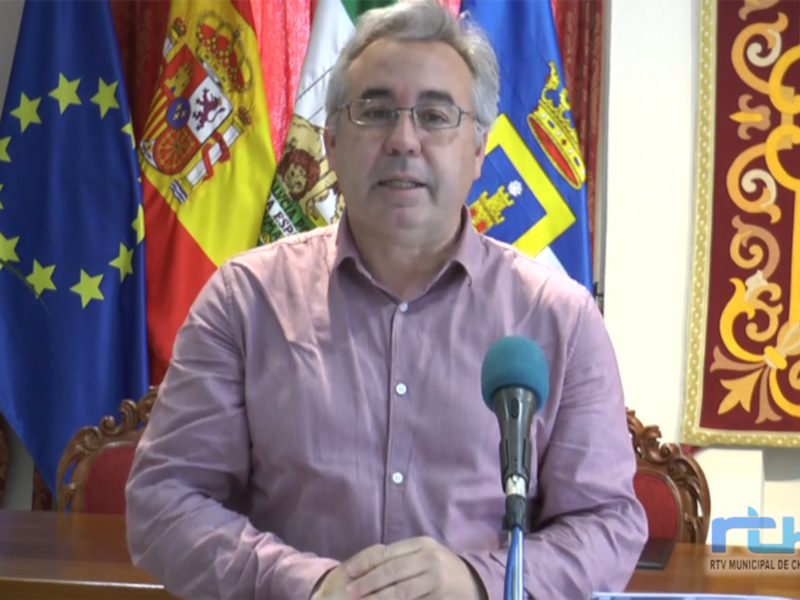 El hombre está sentado en una mesa con un micrófono frente a él. En el fondo, hay varias banderas, incluyendo la de España y la de la Unión Europea. La imagen parece ser de una conferencia o presentación oficial.