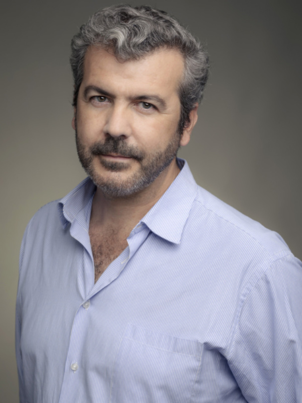 El hombre tiene cabello gris y rizado, barba y bigote. Lleva una camisa azul claro con botones y un bolsillo en el pecho. El fondo es degradado de gris a blanco.