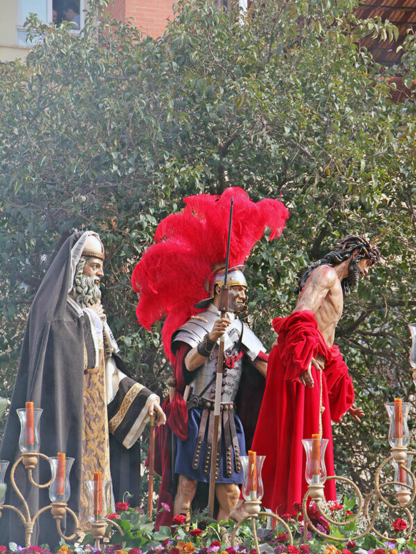Esculturas de San Pedro, un soldado romano y Jesús en la cruz, rodeadas por flores y velas.