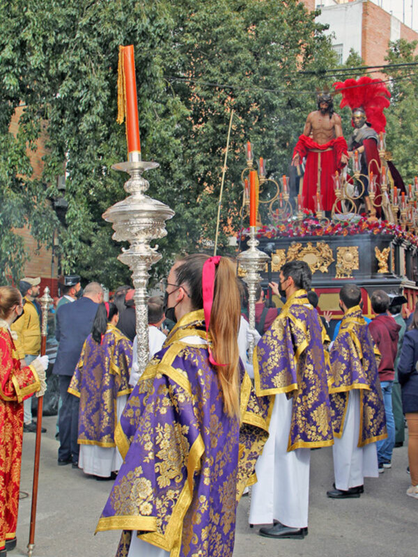 Una procesión religiosa con personas vestidas en trajes tradicionales coloridos, incluyendo una figura central en un carro. La imagen muestra detalles de la vestimenta y el entorno urbano cercano con árboles y edificios en el fondo.