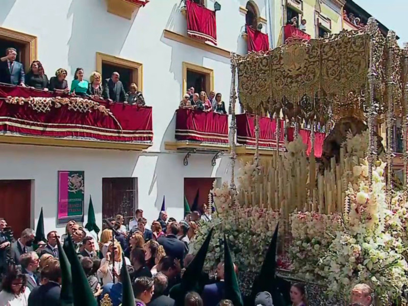 Fotografía de una procesión religiosa en España, con personas en balcones y un carro de flores.