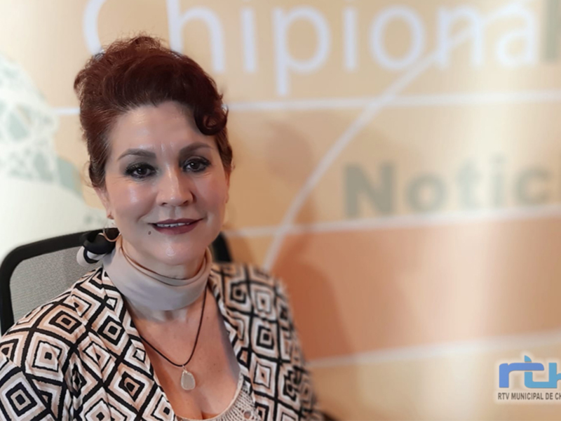 A una mujer con cabello rojo y peinado en un moño. Está vestida con una camisa de manga larga con patrones geométricos en blanco y negro, junto con un collar de cuentas. El fondo es una pared con letras blancas que forman el texto "Chipiona Noticias".