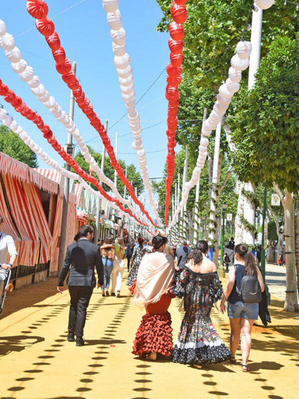 Una pareja camina por un camino decorado con globos rojos y blancos, rodeados de árboles verdes. Algunas personas más caminan a lo largo del camino, disfrutando de la celebración.