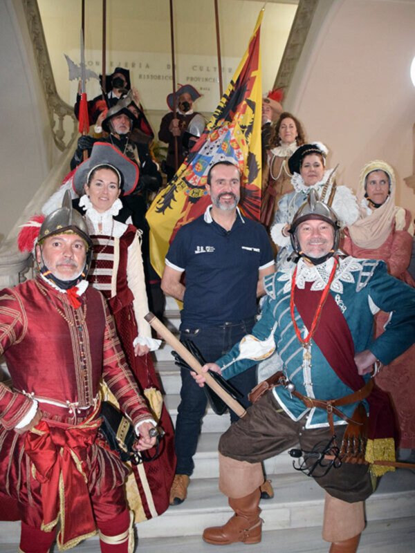 Personas vestidas con trajes históricos posando en una escalera con una bandera en el fondo.