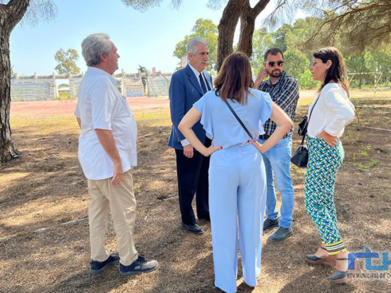 Un grupo de personas conversando en un parque, con una persona en el centro de la imagen, vestida con un traje azul y una corbata, mientras otras personas la escuchan atentamente. El entorno es abierto con árboles y un césped seco.