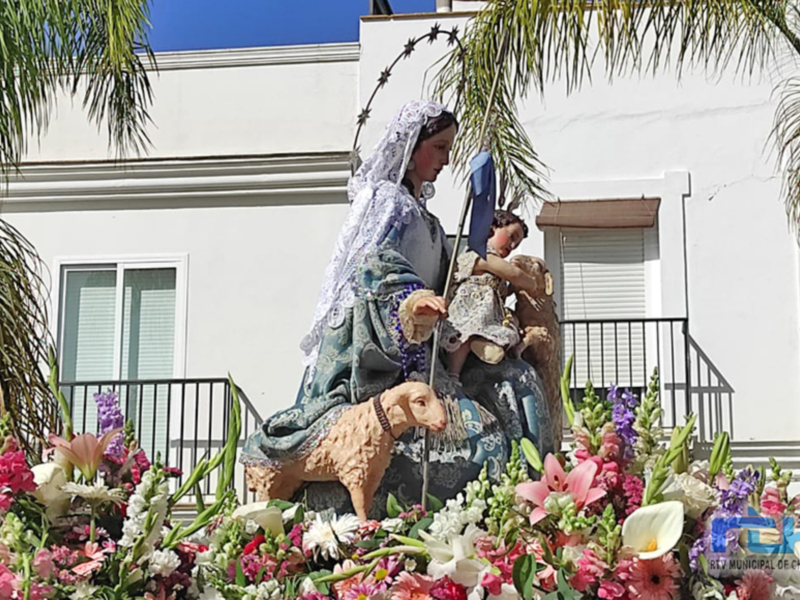 Estatua de la Virgen María con un niño en sus brazos, rodeada de flores coloridas y palmeras. Fondo de una casa blanca con ventanas y un balcón.