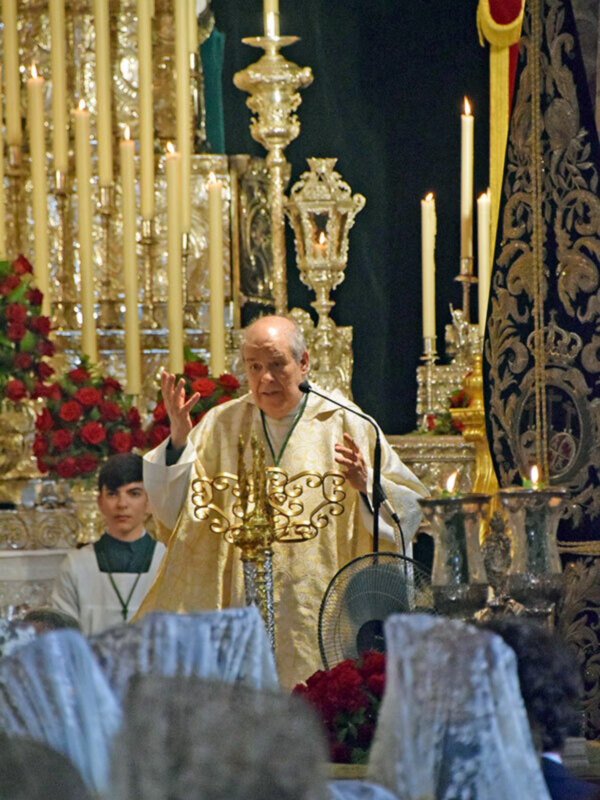 Imagen de un sacerdote en una iglesia, vestido con un hábito dorado y orando frente a una gran candelabro de cristal. El fondo es oscuro, destacándose el resplandor dorado del vestido y la candelabro.