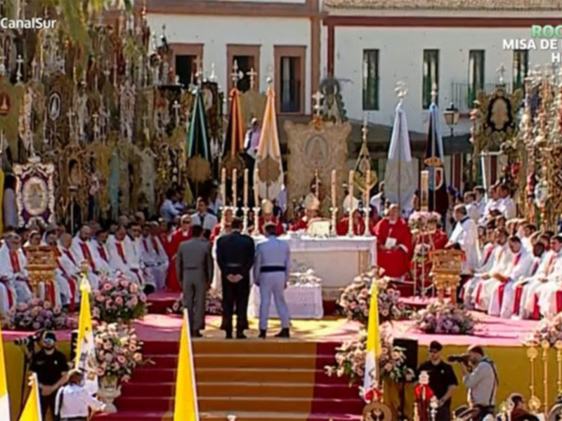 Imagen de una procesión religiosa en un escenario festivo, con personas vestidas formalmente y estatuas o imágenes religiosas en el fondo. La celebración parece ser una misa de romeros, con elementos decorativos y un ambiente festivo.