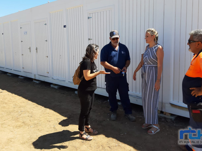Las personas están frente a un edificio de contenedores blancos, discutiendo algo. La escena se da en un área con suelo de tierra y cielo azul claro.