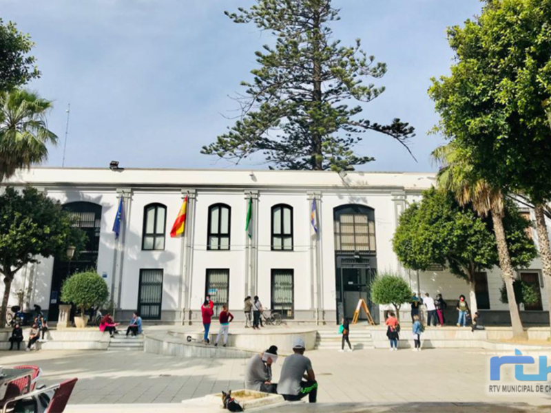 Fachada de un edificio blanco con ventanas y banderas, rodeado por árboles verdes y una plaza con personas sentadas.