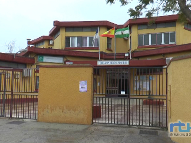 Una escuela pública con un edificio amarillo y rojo. La entrada está protegida por una cerca metálica, y hay varios carteles colgados en las ventanas. En la parte superior de la imagen, se pueden ver banderas españolas ondeando en el viento. La escuela parece ser un lugar de aprendizaje para niños y jóvenes.