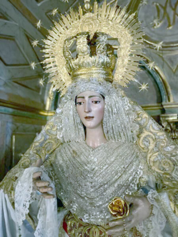 Estatua de la Virgen con vestimenta dorada y blanca, coronada por una corona estilizada. Contexto: interior de una iglesia con detalles artísticos y arquitectónicos.