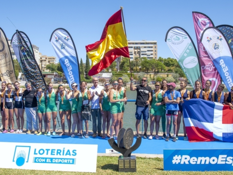 Equipo de remo español celebrando una victoria junto a banderas y patrocinadores.