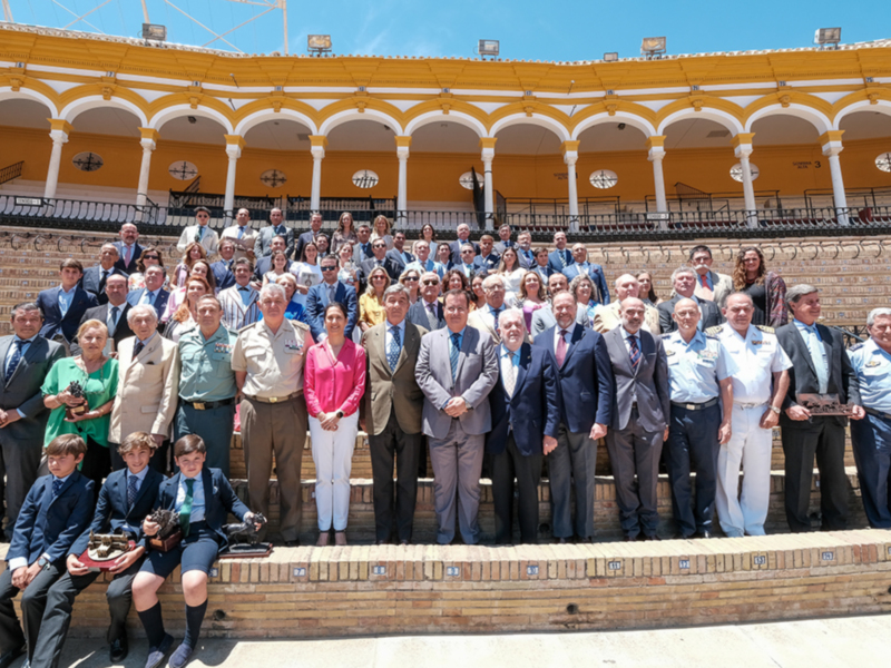 Grupo de personas posando en el interior de una plaza de toros, con palcos y estructuras de piedra al fondo.