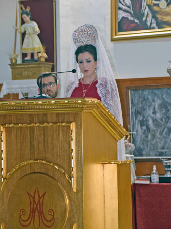 Una mujer con un vestido blanco y una capa de encaje se dirige a un público. En el fondo, hay pinturas religiosas y una mesa con una botella de agua.