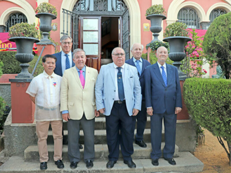 Seis hombres en trajes formales posan juntos en un patio con jardín y edificio de fondo.