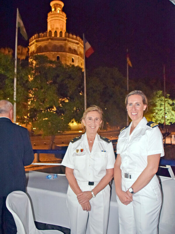 Dos mujeres en uniforme naval posan sonrientes en una cena formal, con la Torre del Oro de la ciudad de Sevilla al fondo.