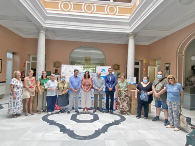 Una imagen de una reunión formal en un edificio histórico, con personas vestidas formalmente y una mesa con productos en el centro. La decoración incluye columnas y una gran puerta al fondo.