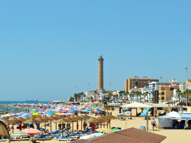 Playa con sombrillas, toldos y una torre en el fondo bajo un cielo azul.