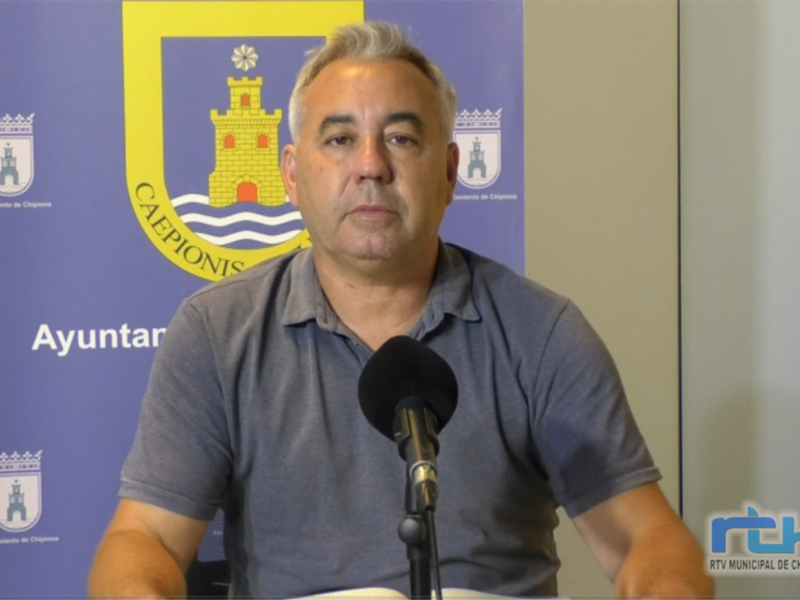 El hombre está en una conferencia de prensa frente a un fondo con el escudo y la bandera de Chipiona. Se puede leer "Ayuntamiento" en la pared. La imagen es de RTVC y muestra a una persona hablando por un micrófono en una mesa.