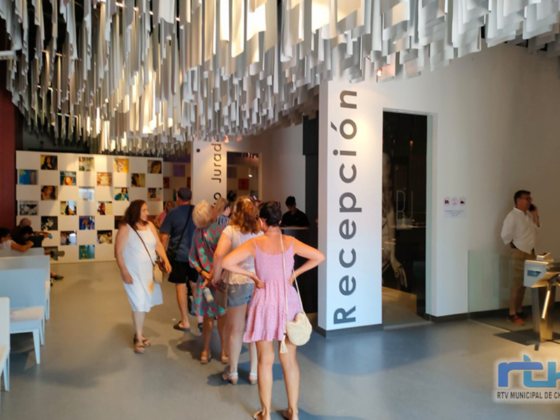 Recepción en un edificio moderno con una estructura de paneles blancos y colgantes. Personas caminando por el pasillo, mostrando un ambiente de visita o exposición.