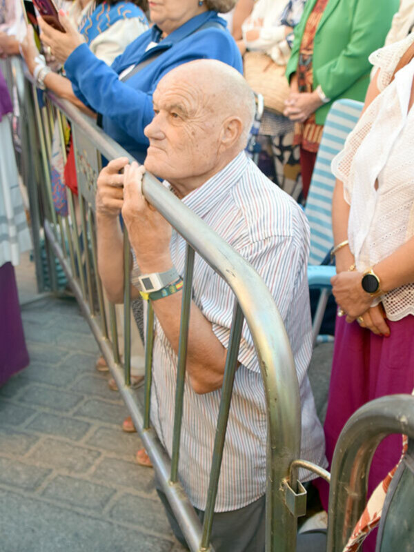 Un anciano con camisa a rayas y pantalones va a la parada de un tranvía. Detrás hay personas con vestimenta casual y algunos con bolsos. La imagen muestra una escena de transporte público en un entorno urbano.