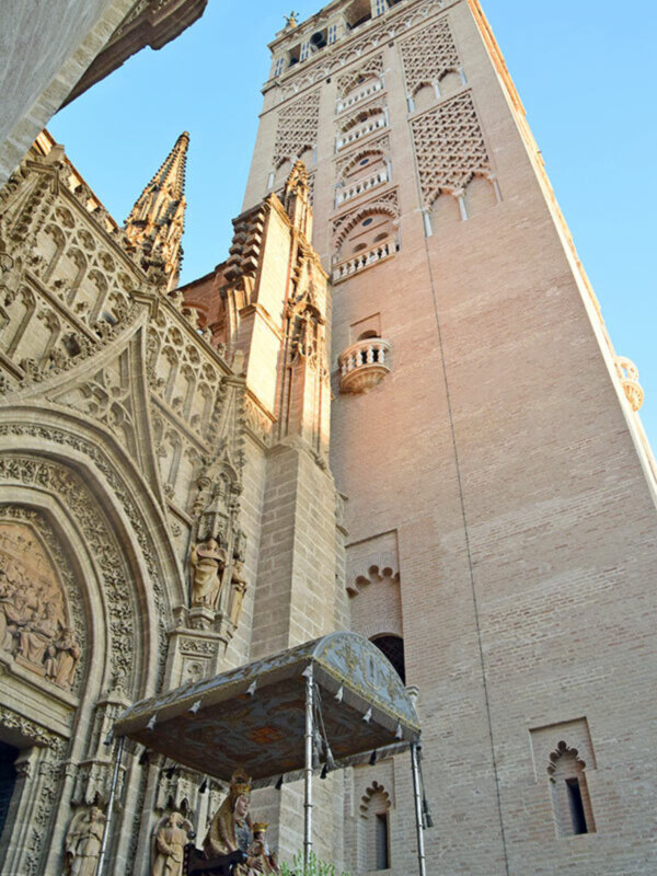 Una vista de arriba hacia abajo de la torre de una mezquita, con detalles geométricos y decorativos en el exterior. La torre es alta y tiene una estructura compleja con ventanas y molduras. En la base de la imagen, se ve una estructura metálica con un toldo, probablemente para proteger a los visitantes. El cielo azul claro se puede ver en el fondo, iluminando la estructura de la mezquita.