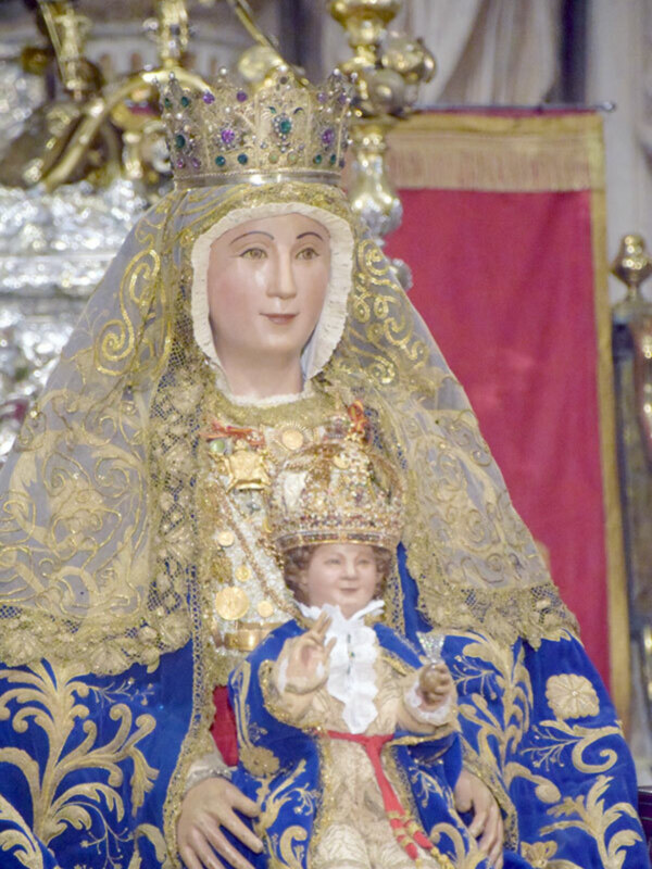 Imagen de la Virgen del Rocío, vestida con traje azul y oro, y el Niño Jesús en su regazo, rodeada de ornamentos y joyas.