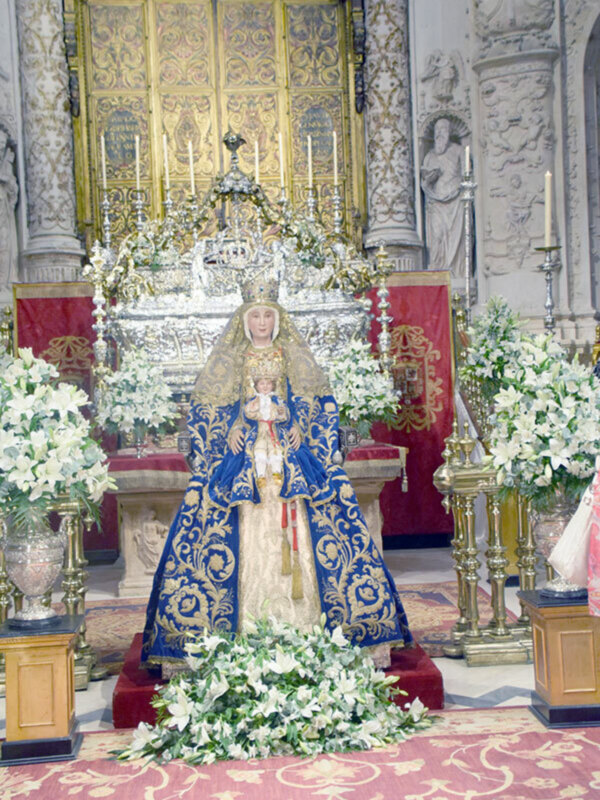 Imagen de la Virgen María en un trono ornado con flores y velas, en un entorno de iglesia con detalles dorados y ornamentación.