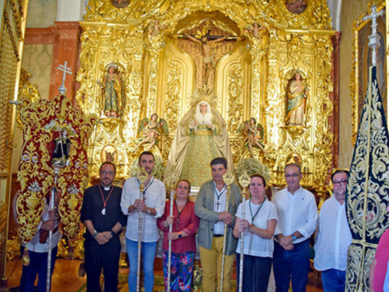 Una procesión religiosa con una imagen de la Virgen María en el centro, rodeada de personas vestidas formalmente y portando estandartes. La imagen muestra una escena festiva y ceremonial en un entorno religioso.