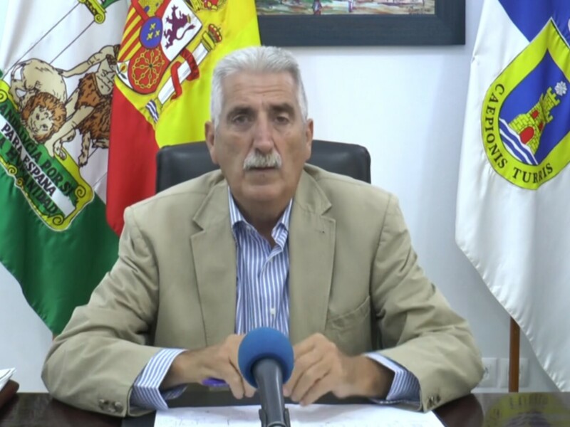 El pié de foto es un hombre sentado en una silla, con una bandera española y otra nacional detrás de él. Está hablando en un micrófono, con documentos sobre la mesa delante.