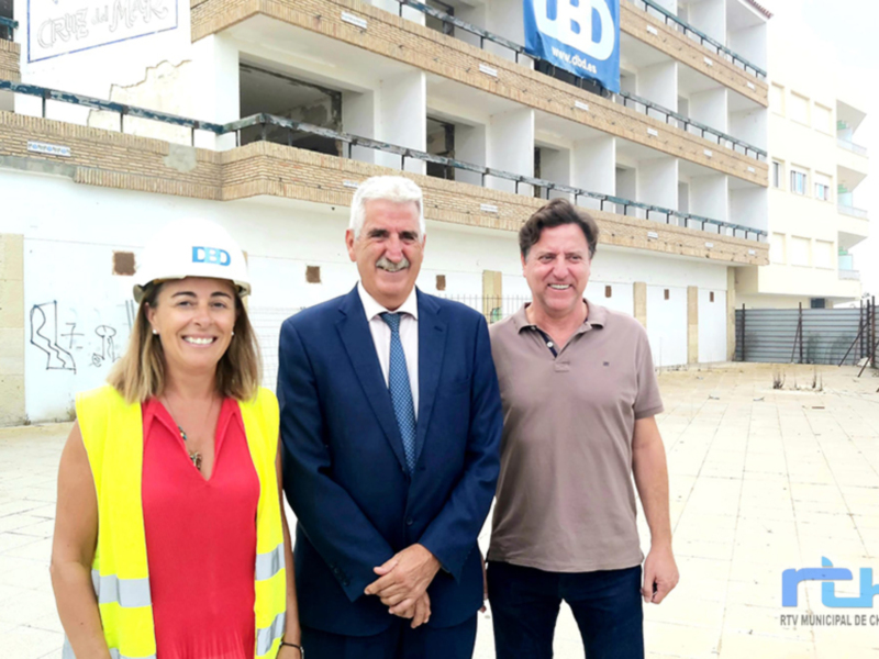 En construcción, tres personas posan frente a un edificio en fase de desarrollo. La imagen muestra una estructura urbana con varios niveles y ventanas, destacando la construcción en progreso. La escena transmite el avance de un proyecto inmobiliario, con una persona en casco y otro en traje formal.