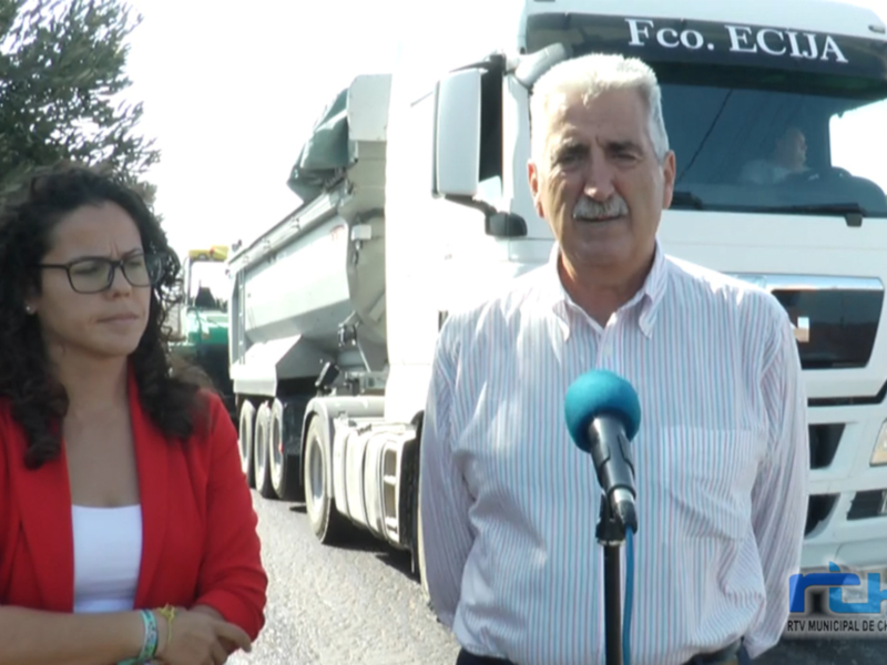 Dos personas, una mujer y un hombre, están en el lado izquierdo de la imagen. La mujer está hablando al microfono mientras el hombre, vestido con una camisa blanca y azul, la mira. Al fondo hay un camión de carga blanco con el nombre "Fco. ECIJA" en la parte superior derecha. La escena parece ser al aire libre, posiblemente en una zona industrial o de transporte.