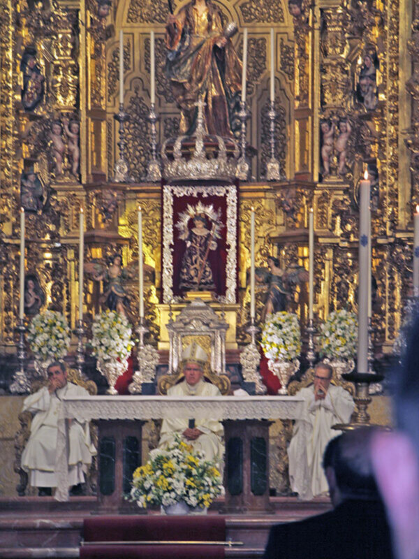 Imagen de una iglesia con altares dorados, estatuas y flores. Personajes vestidos de blanco en el altar.