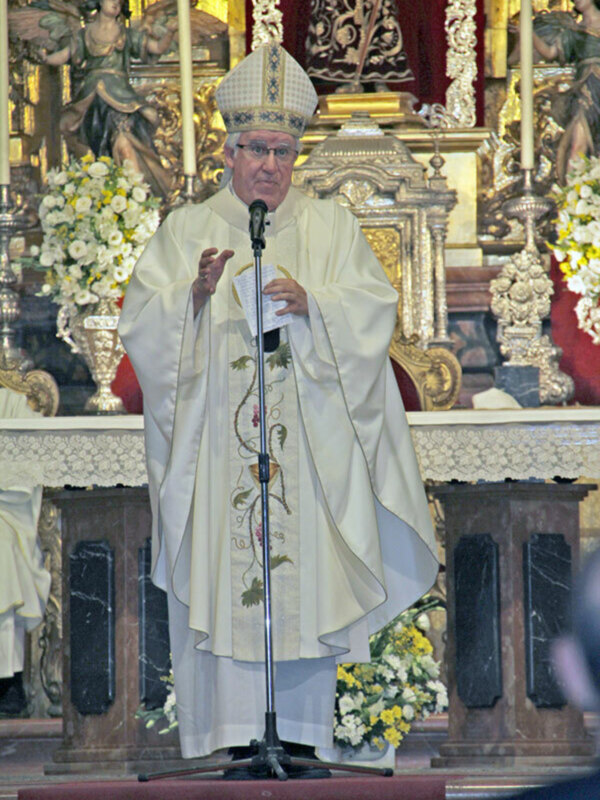 Un sacerdote en vestimenta bíblica se encuentra frente a un altar con flores blancas y amarillas, mientras pronuncia una oración. La imagen muestra detalles de un altar con adornos dorados y figuras religiosas en el fondo.