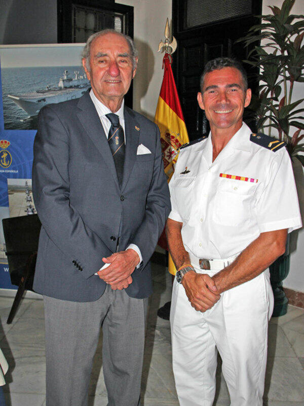 Dos hombres, uno en traje y el otro en uniforme naval, se saludan en una sala con una bandera española y un póster de un barco en el fondo.