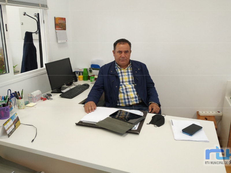 Un hombre está sentado en un escritorio, trabajando con documentos y una computadora. La imagen muestra detalles de su entorno laboral, incluyendo un teléfono móvil y una agenda. La imagen también muestra un espejo en el fondo, que refleja la habitación.