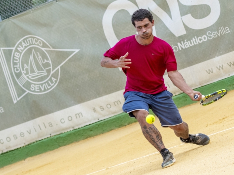 Jugador de tenis en acción en el Club Náutico Sevilla, con un fondo que incluye el logotipo del club y un banner con información sobre el evento.