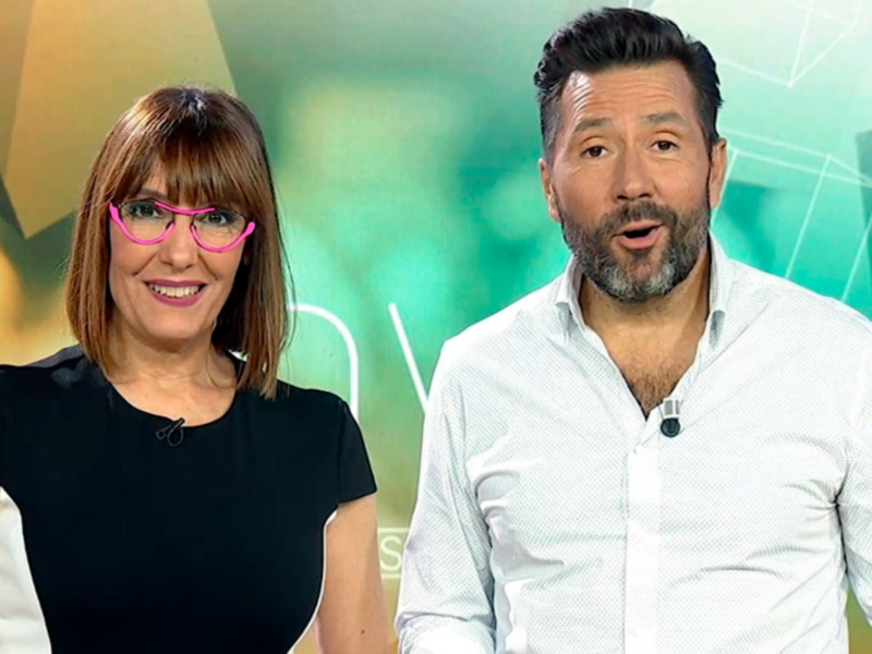 Dos presentadores en un estudio de televisión con un fondo verde y amarillo. La mujer lleva una camiseta negra y gafas de color rosa, mientras que el hombre está vestido con una camisa blanca.