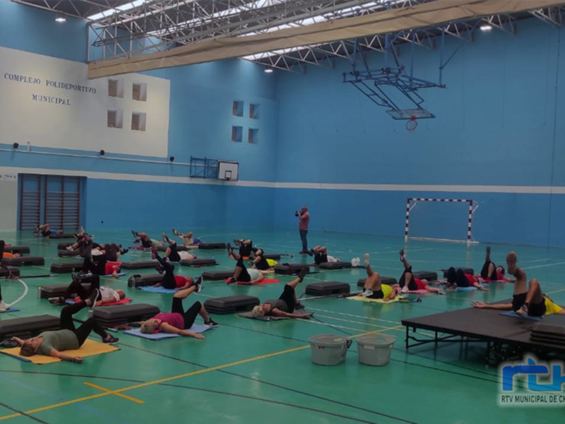 En una gran sala de deportes, un grupo de personas realiza ejercicios en colchonetas. Se observa a una persona tomando notas o grabando la actividad, mientras otros participantes realizan ejercicios de estiramiento y flexibilidad. La sala está iluminada con luces blancas, y en el fondo se puede ver una cancha de baloncesto.