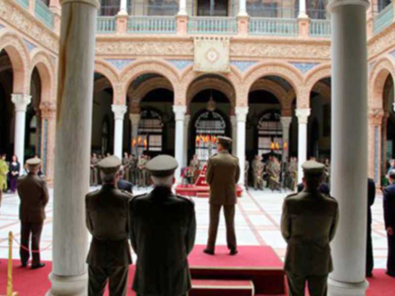 Imagen de un palacio real con columnas y arcos, donde varios oficiales militares se encuentran en un acto formal. El ambiente es serio y ceremonial, reflejando la importancia del evento.