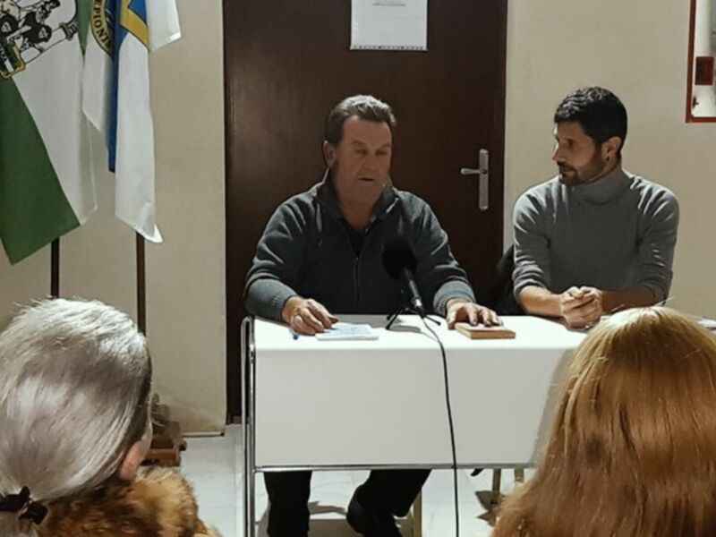 Conferencia de prensa con dos hombres sentados en una mesa con banderas y un micrófono en el centro.