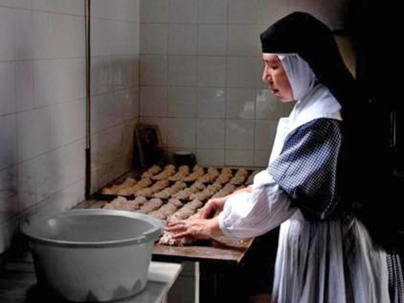 En una cocina con azulejos blancos, una monja con un hábito negro y blanco prepara pan en una mesa de madera. A su lado, un recipiente de cerámica está listo para la próxima tarea.