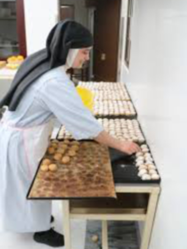 Una monja en un hábito blanco y negro está preparando panes en una cocina. La imagen muestra una mesa con varios huevos, un huevo cocido y una bandeja de panes. La monja está sentada frente a la mesa, con su rostro visible y una expresión concentrada. La cocina tiene un estilo tradicional con armarios de madera y una puerta abierta al fondo.