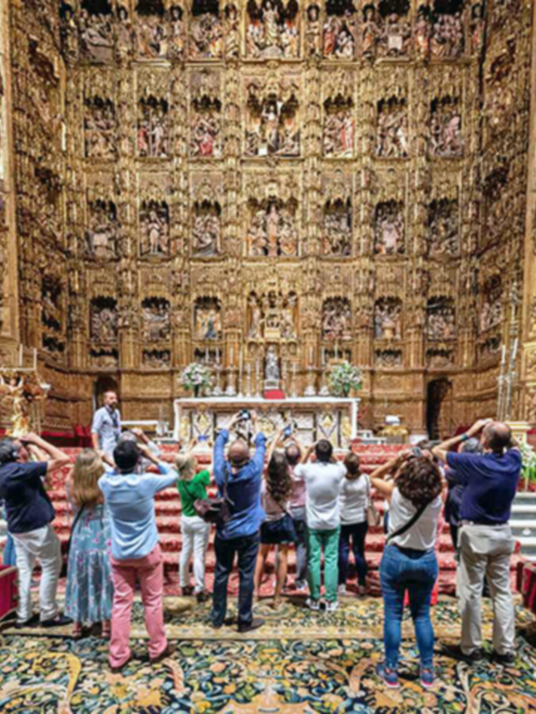 En una majestuosa iglesia, un grupo de personas levanta sus manos en un gesto de adoración. El interior está adornado con una impresionante pared de nichos llenos de esculturas religiosas, mientras que el altar central destaca con flores y una cruz. La arquitectura dorada y la alfombra de colores vibrantes crean un ambiente de reverencia y devoción.