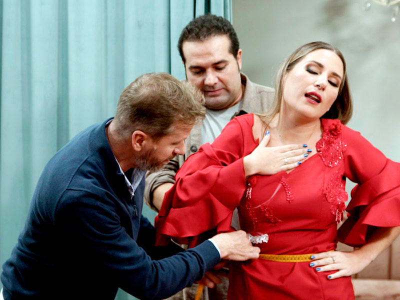 Una mujer en un vestido rojo con detalles bordados se muestra a una persona que la ayuda a ajustar su cintura con un cinturón dorado. La escena ocurre en un entorno interior, posiblemente una tienda de modas o un salón de belleza.