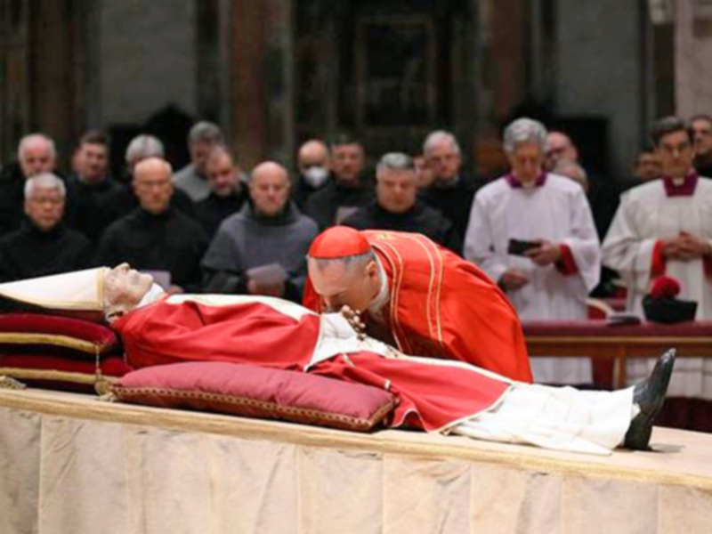 Imagen de un cardenal en una iglesia, con una saya roja y un sombrero de papa, mientras lee un libro.